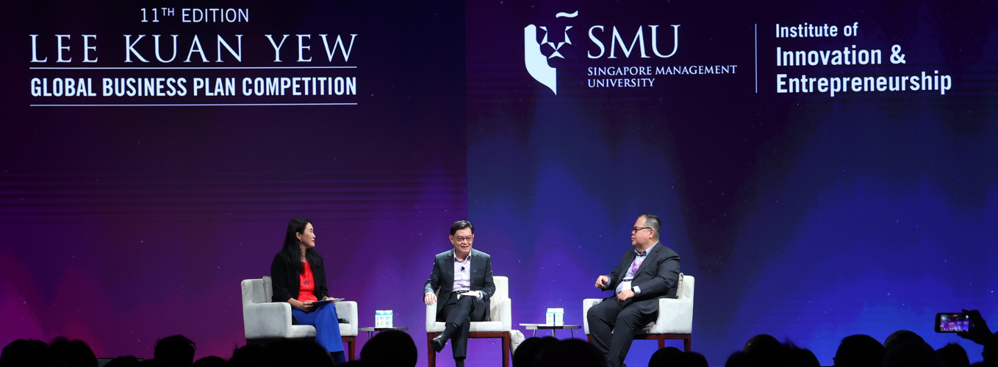 Three people seated on stage at the Lee Kuan Yew Global Business Plan Competition; audience visible.